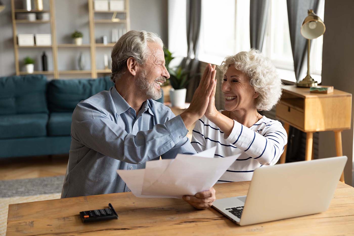 Overjoyed older married couple giving high five after finishing paperwork together at home.