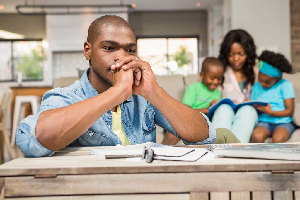 Father sits over bills with concerned expression, his happy family sitting together in the background.